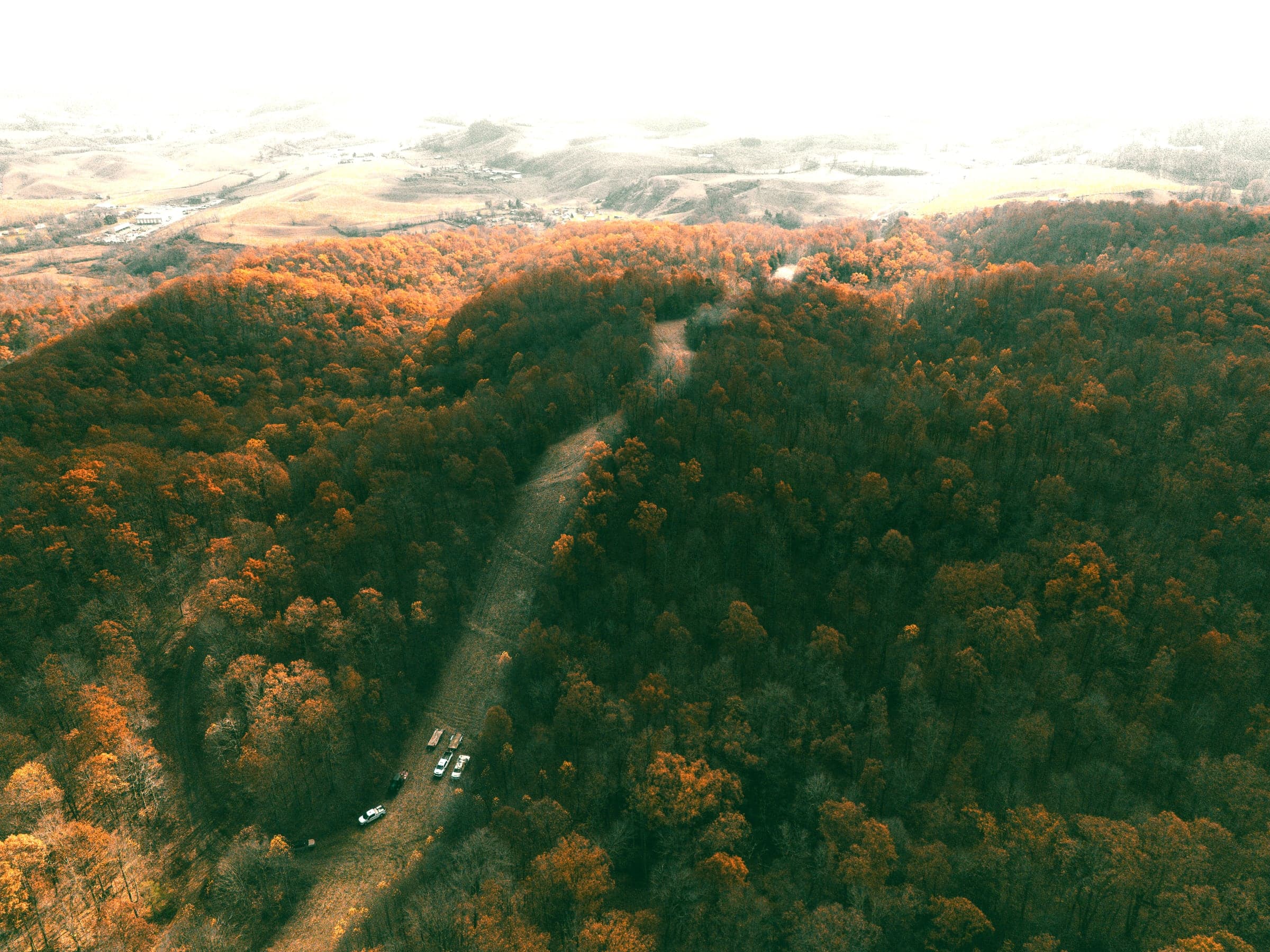 Aerial view of a right of way cut running through mixed hardwood forest in autumn, with maintenance trucks and trailers staged in the cleared strip.