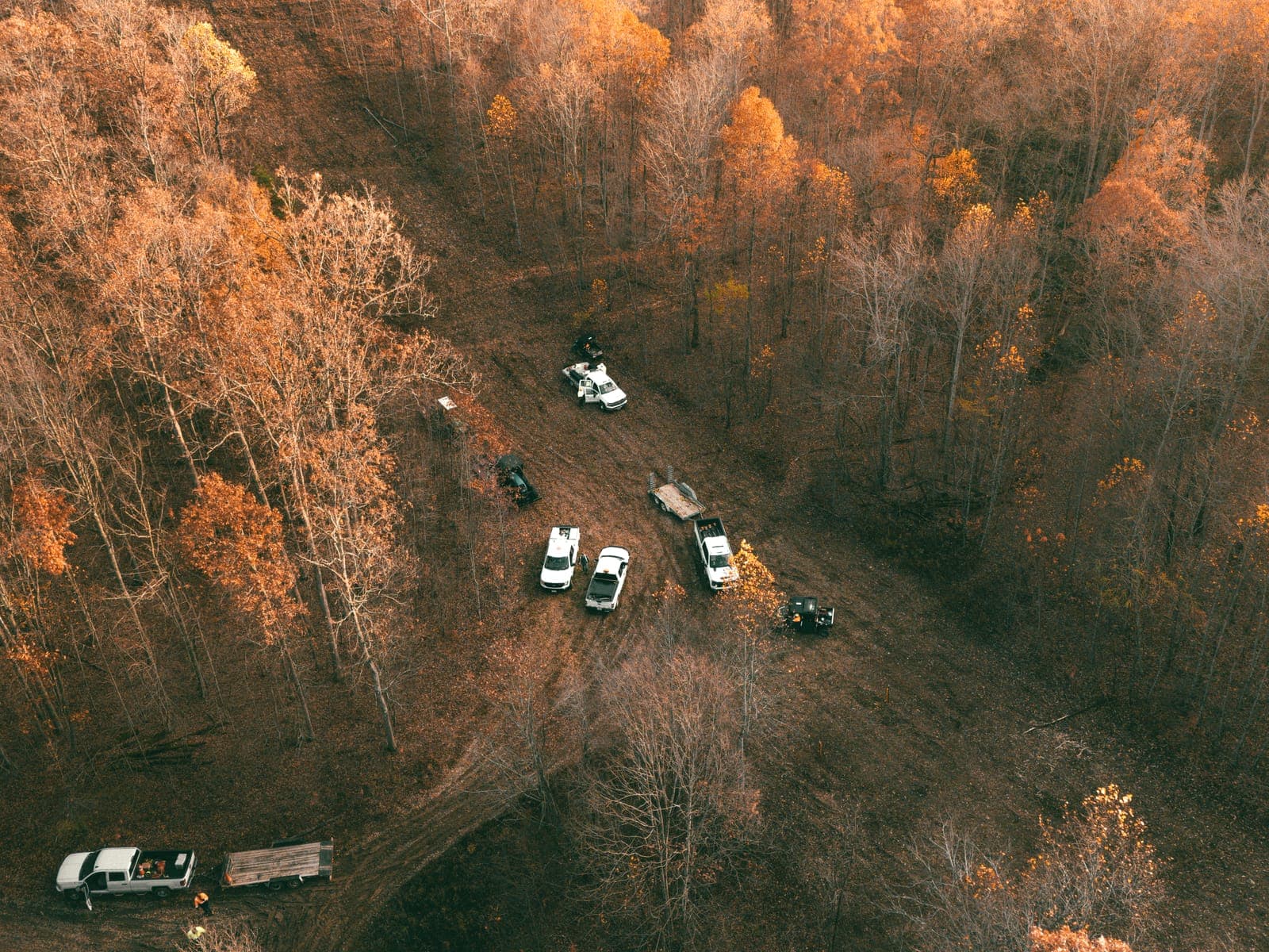 Aerial view of trucks and trailers staged along a right of way in late autumn, with bare hardwood on either side.
