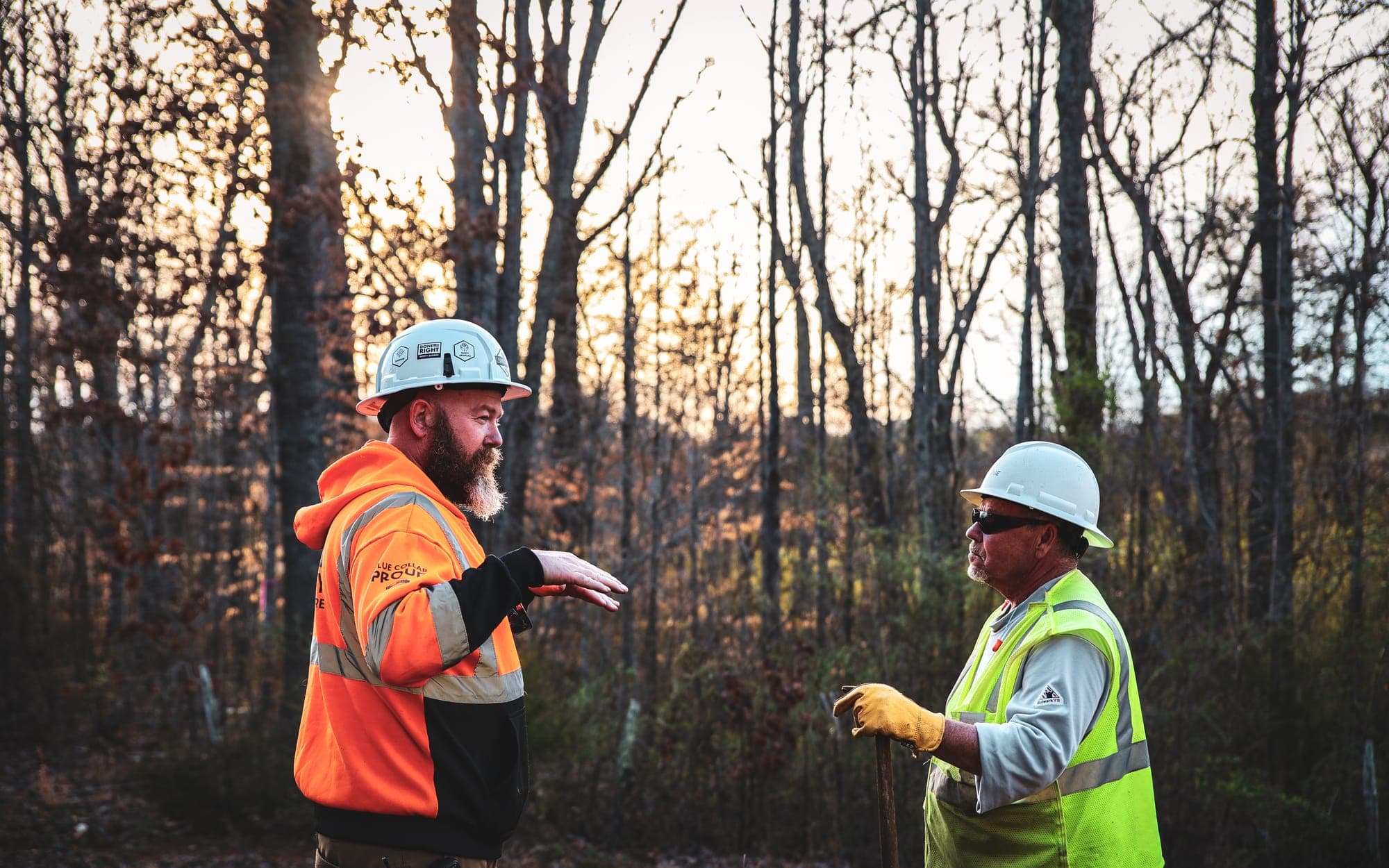 Two vegetation management crew members in high-visibility clothing and hard hats discussing a stretch of right of way in open woodland.